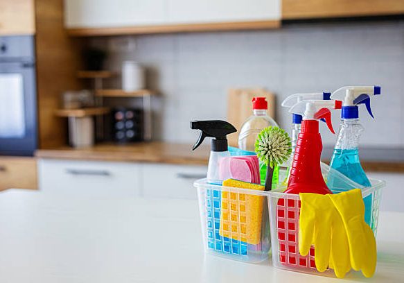 A plastic basket filled with various cleaning supplies including spray bottles, sponges, rubber gloves, and brushes placed on a white kitchen countertop. The background shows a modern kitchen with wooden and white cabinets, perfect for illustrating home cleaning, hygiene, or housekeeping concepts.